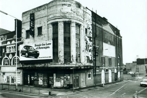 Black and white image of the outside of a grand building on a corner. The building looks run down and closed.
